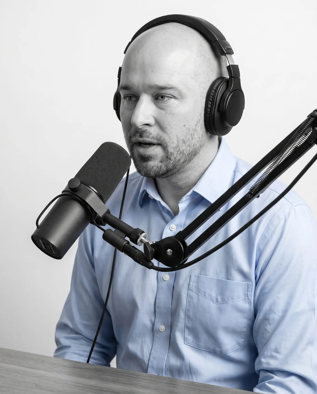 Ryan in a light blue shirt and black over-ear headphones, speaking into a studio microphone on a desk arm, in black and white with only the shirt in colour